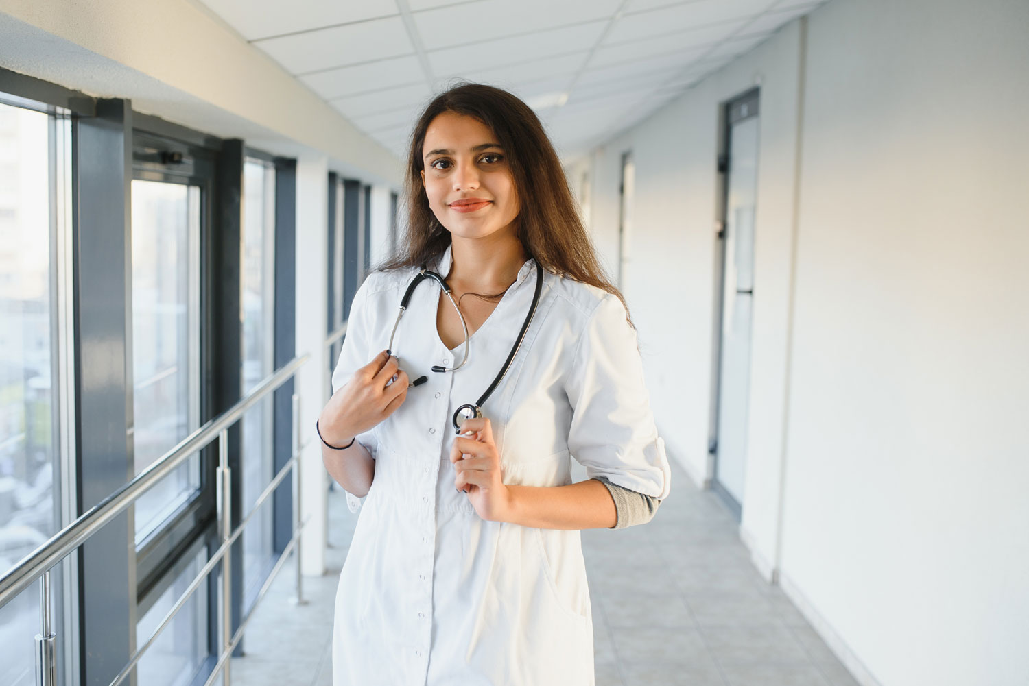doc1 A person wearing a white lab coat and a stethoscope stands in a hallway with large windows, smiling at the camera.